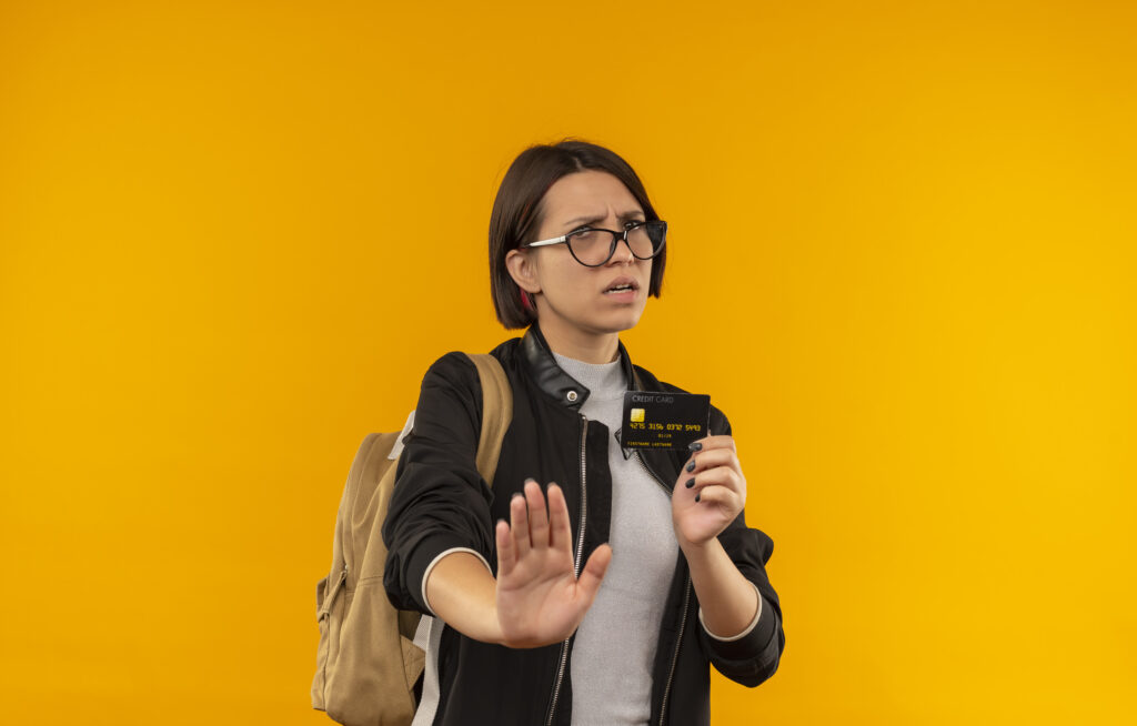 unpleased young student girl wearing glasses back bag holding credit card gesturing stop isolated orange background with copy space