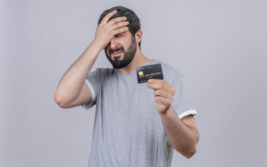 stressed young handsome man holding credit card putting hand head with closed eyes isolated white wall